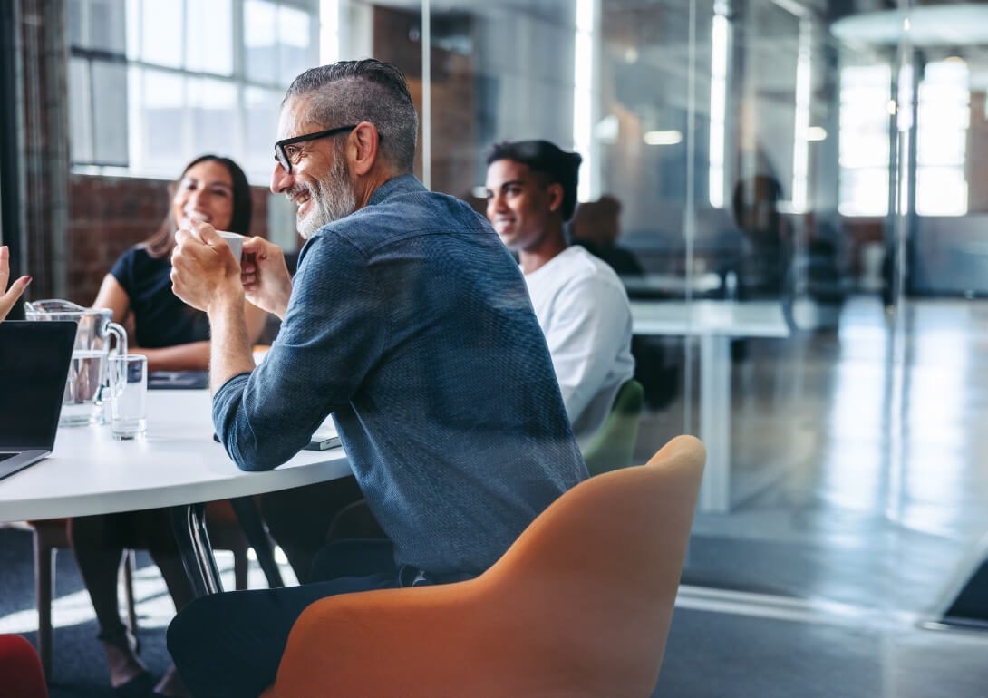 A seasoned leader with grey hair and glasses is captured from the back, enthusiastically engaging with his diverse team during a training session. They are seated around a modern conference table in a bright, open-plan office space. The team looks on, their attention captured by the discussion, symbolizing the leader-led delivery model where internal leaders utilize playbooks to guide learning within the company.