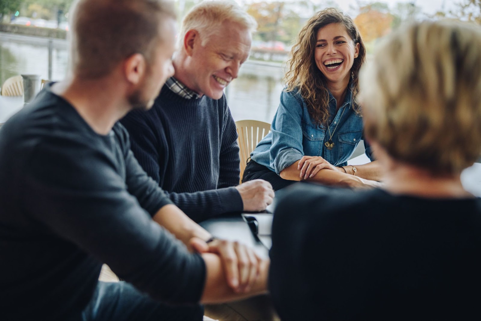 A lively group of professionals shares a light-hearted moment around a table, with one woman laughing joyously in the center of the conversation. The atmosphere suggests a sense of camaraderie and inclusion, essential for a consistent learning experience that values each participant and ensures the retention of key messages within the team.